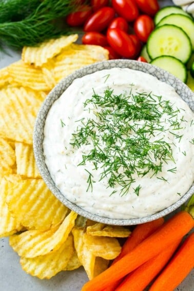 A bowl of creamy dill dip surrounded by potato chips and vegetables.