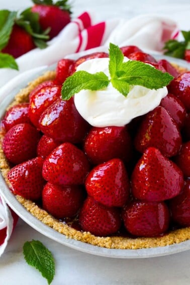 A pile of strawberries inside a graham cracker crust, topped with a shiny red glaze, whipped cream and mint.