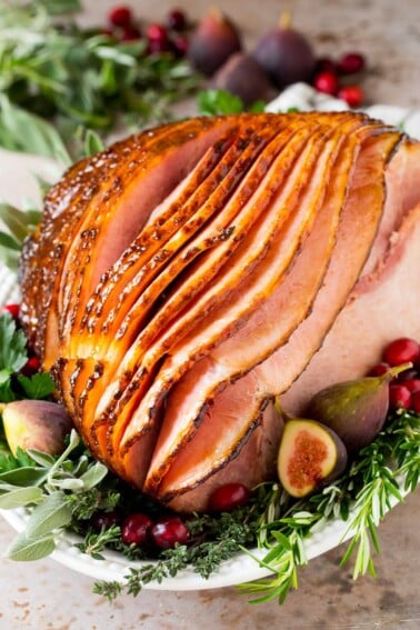 A honey glazed ham on a serving platter garnished with herbs and fruit.